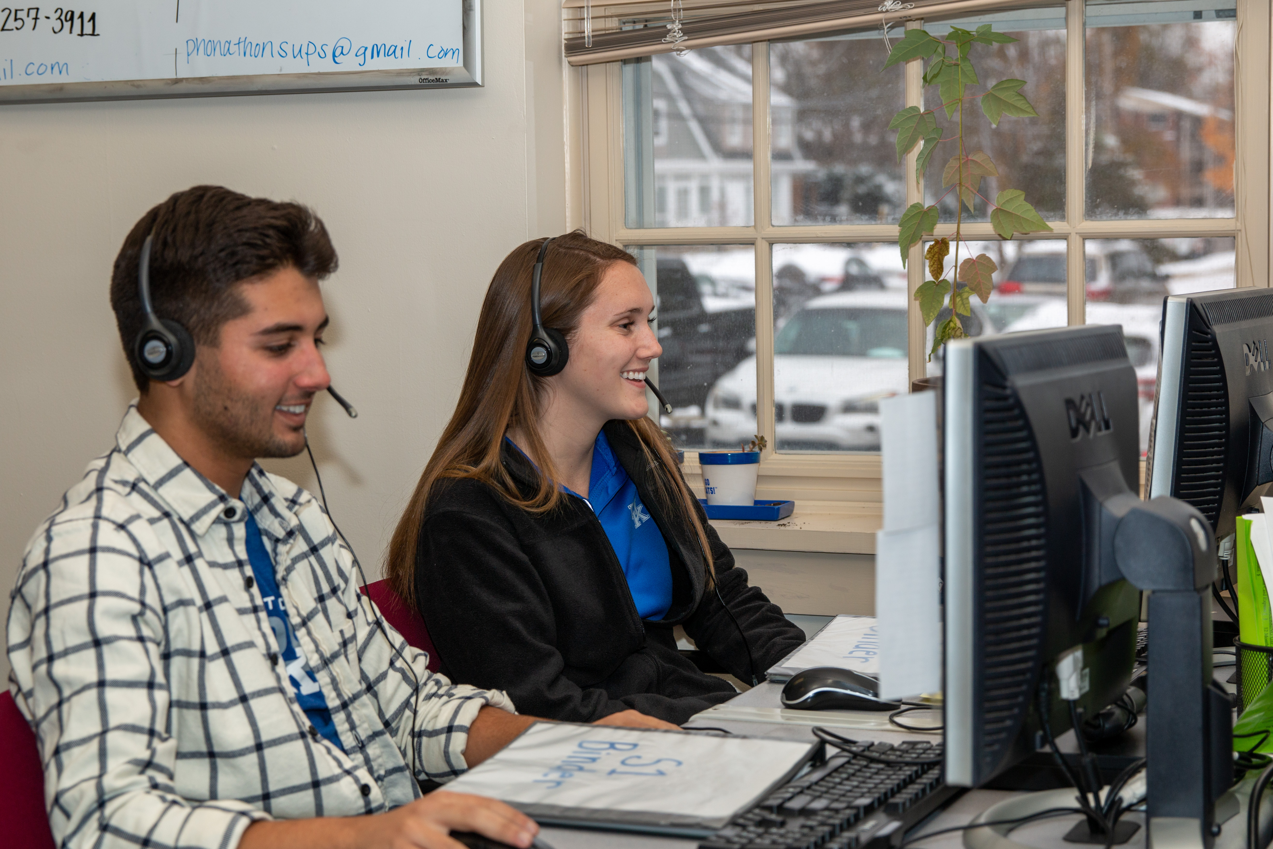 Two students are seated in front of computers, laughing and wearing headsets